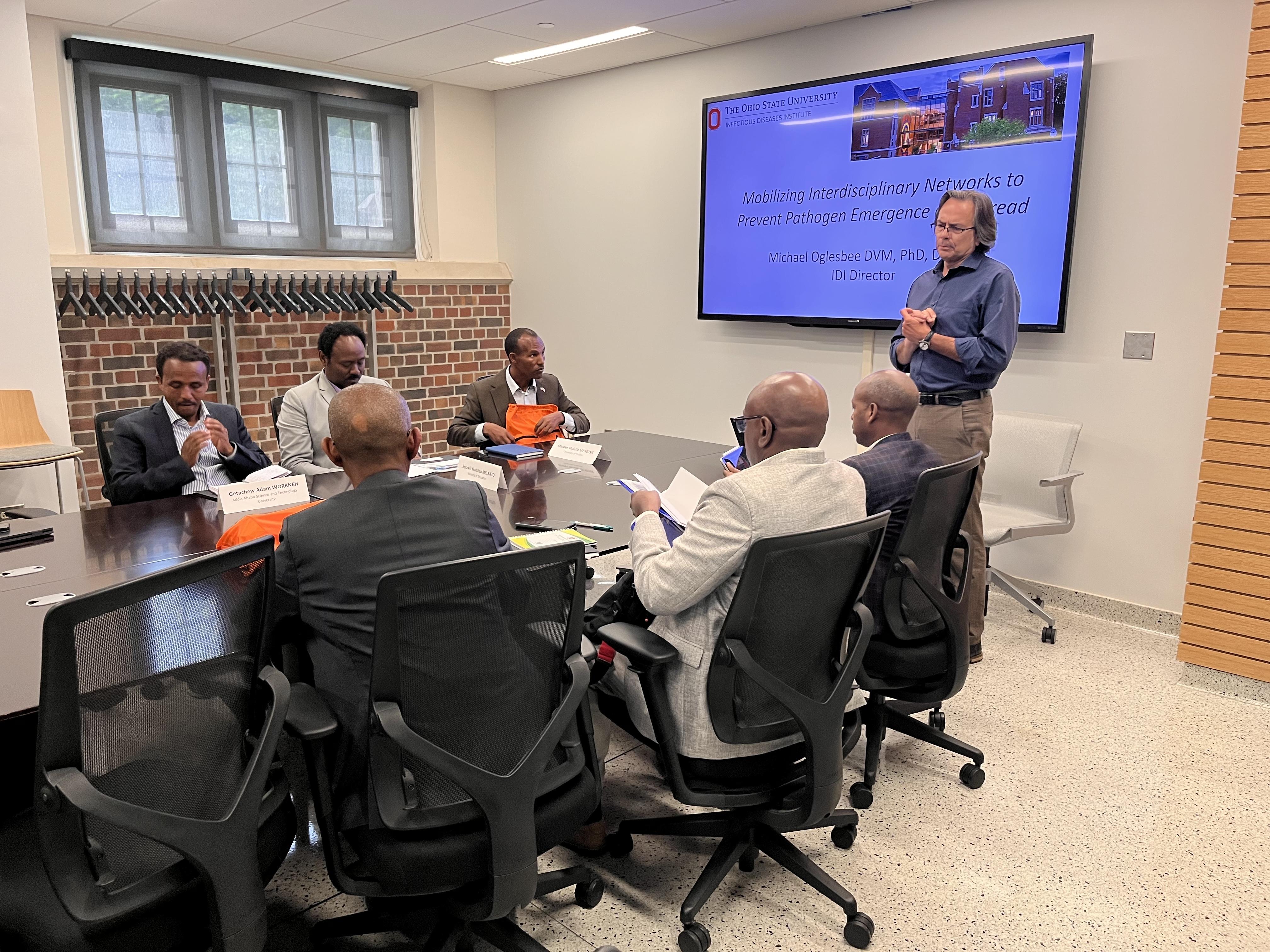 Group of people seated around a conference table in a meeting room, viewing a presentation on a large wall-mounted screen.