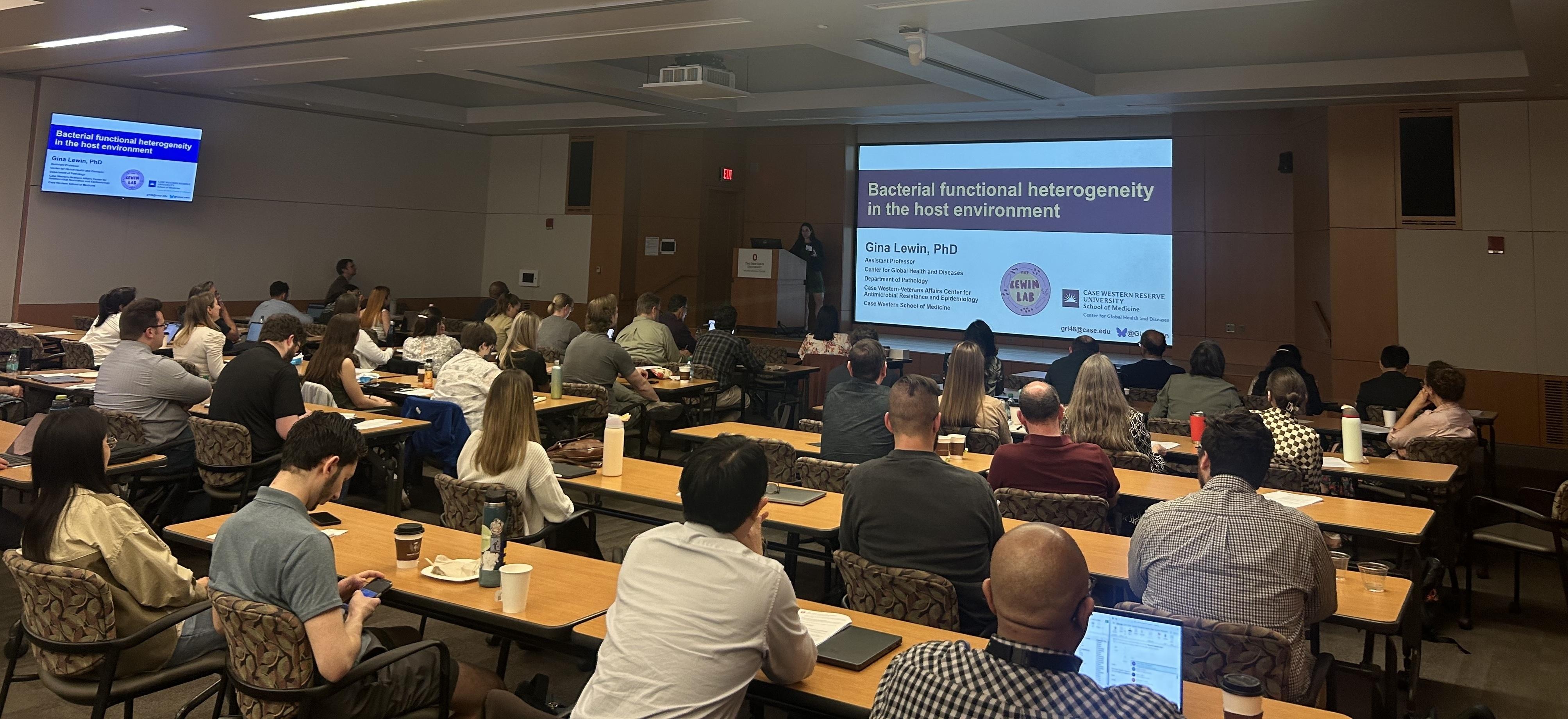Conference room with attendees seated at tables, watching a presentation titled 'Bacterial functional heterogeneity in the host environment' by Gina Lewin, PhD, displayed on a large screen.