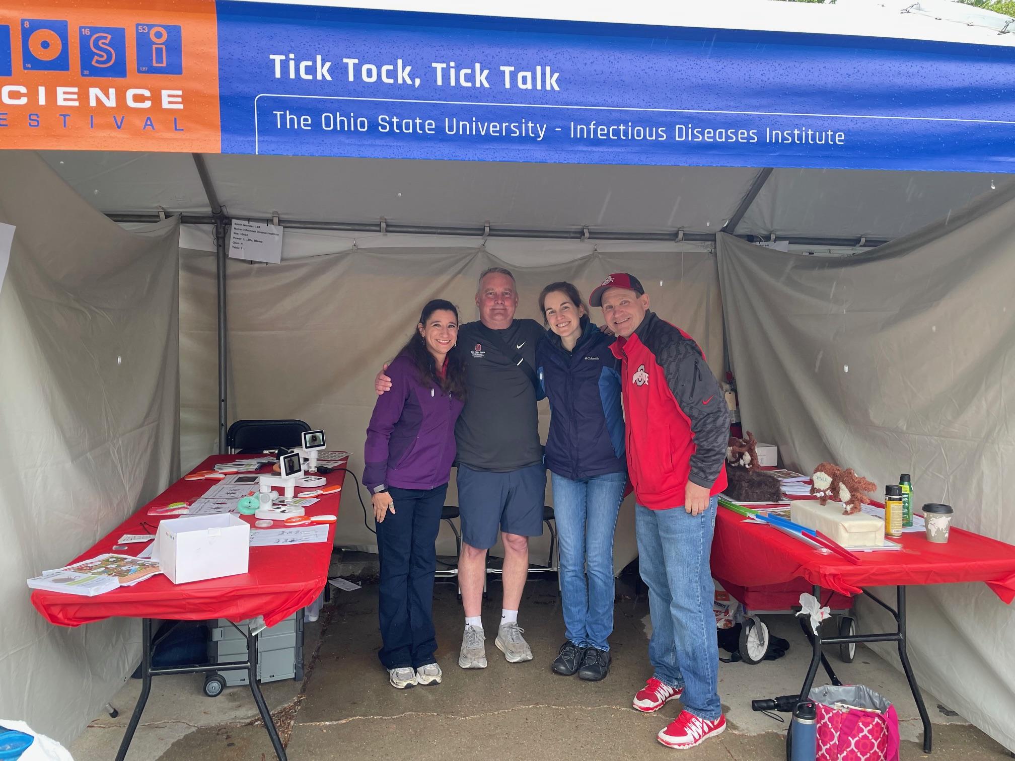 Booth at the COSI Science Festival featuring a banner that reads 'Tick Tock, Tick Talk' and 'The Ohio State University - Infectious Diseases Institute,' with four individuals posing under the tent and tables displaying educational materials.