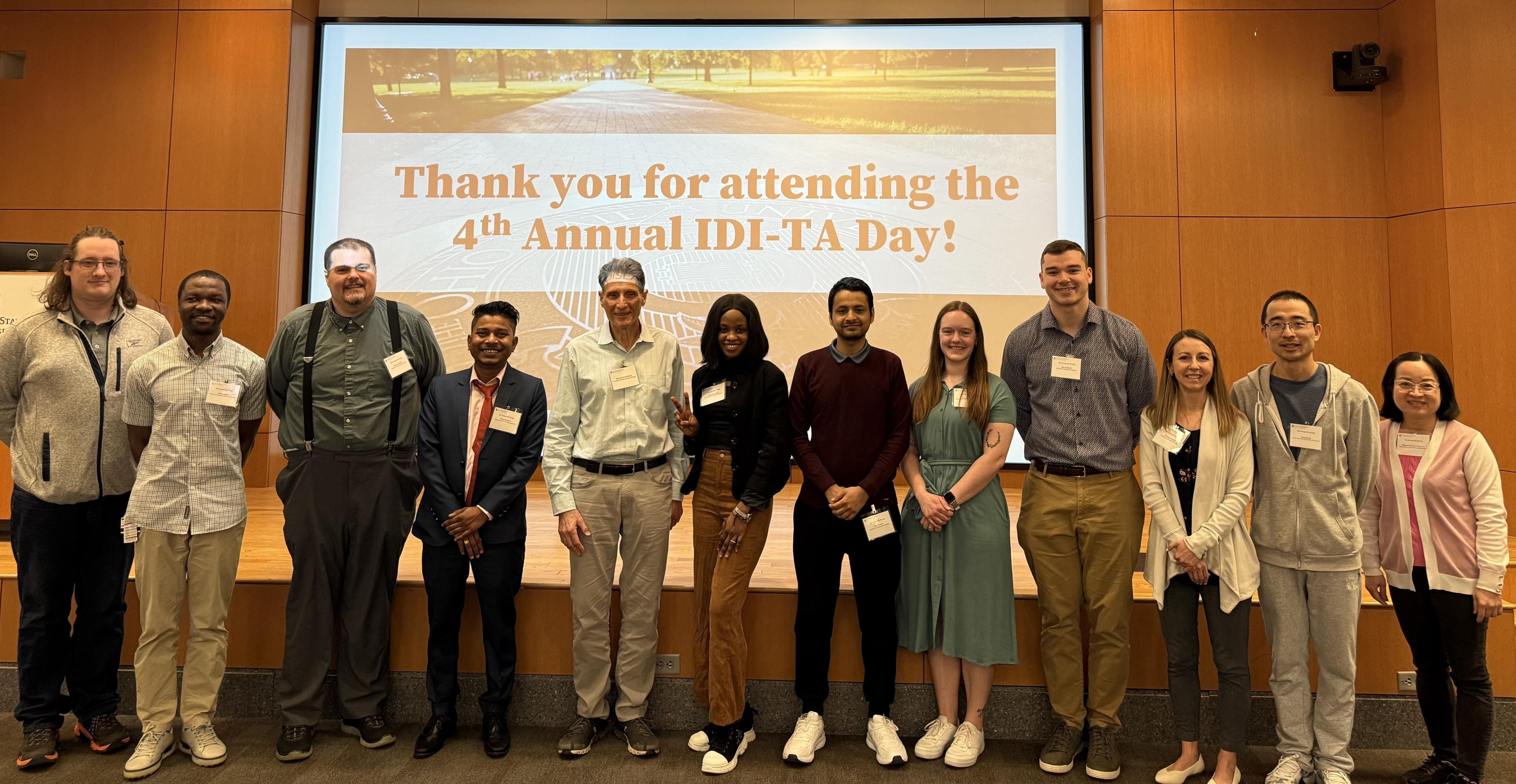 Twelve individuals standing in front of a large screen displaying 'Thank you for attending the 4th Annual IDI-TA Day!' in a room with wooden paneled walls; group includes a mix of casual and formal attire.