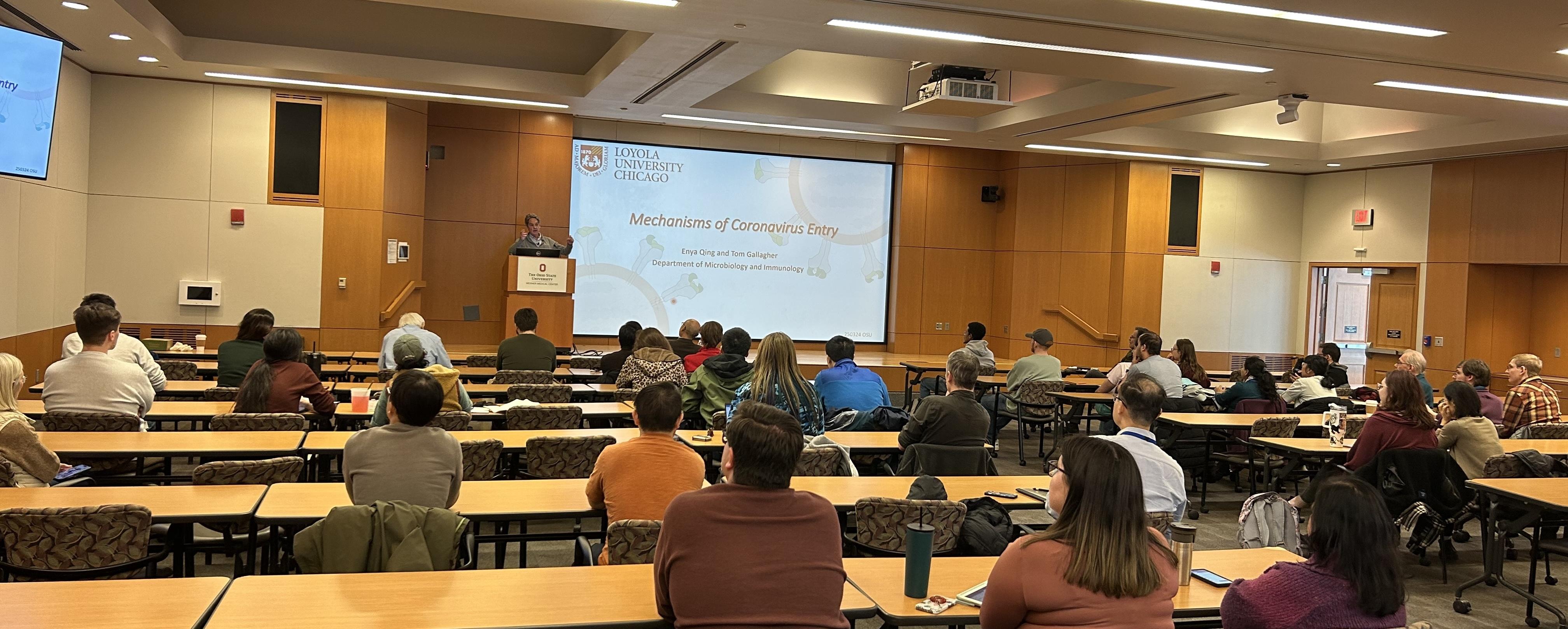 Lecture hall with attendees seated at tables, listening to a presentation titled 'Mechanisms of Coronavirus Entry' by a speaker at a podium. The screen displays equations related to SARS-CoV-2 and the Loyola University Chicago logo.