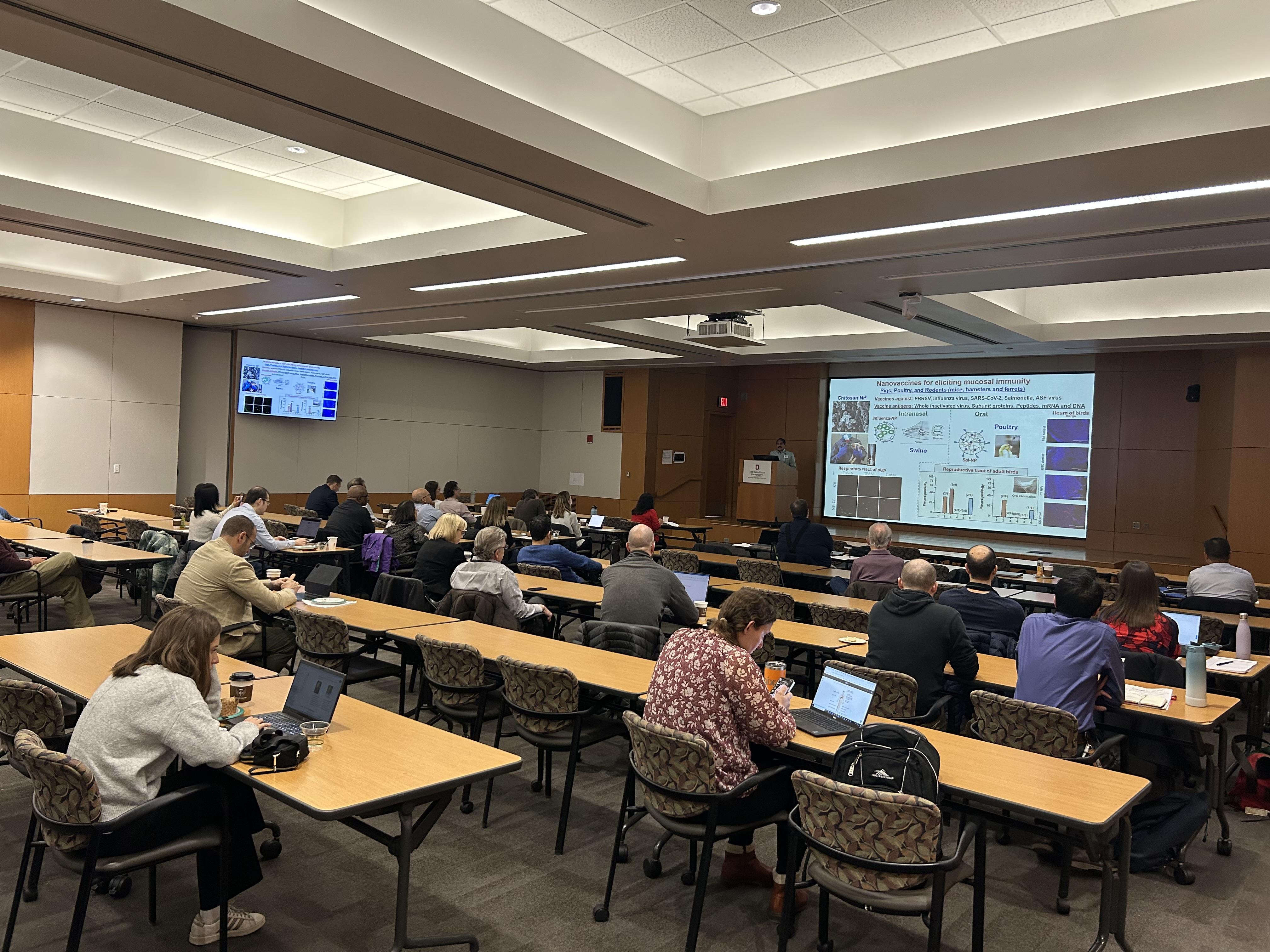 Audience seated in a conference room listening to a presenter with slides projected at the front.