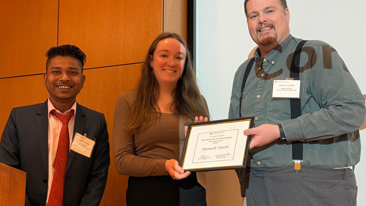Three individuals stand together, with the person in the center holding a framed certificate. The person on the left wears a suit and tie, the center person wears a long-sleeved shirt, and the person on the right wears suspenders over a button-up shirt.