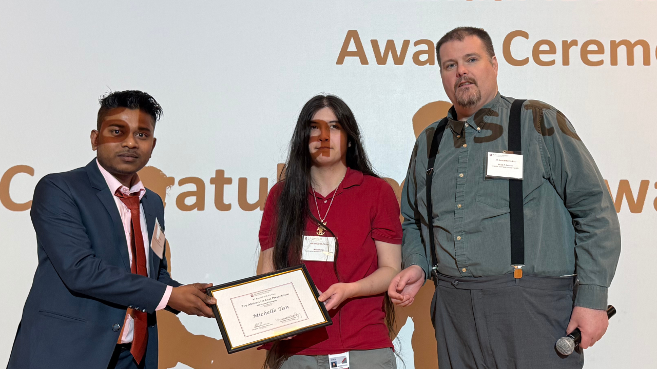 Three individuals at an award ceremony for the 4th Annual IDI-TA. One person hands a certificate to the recipient in the center, while another holds a microphone. A backdrop behind them displays the words “4th Annual IDI-TA Award Ceremony” and “Congratulations.