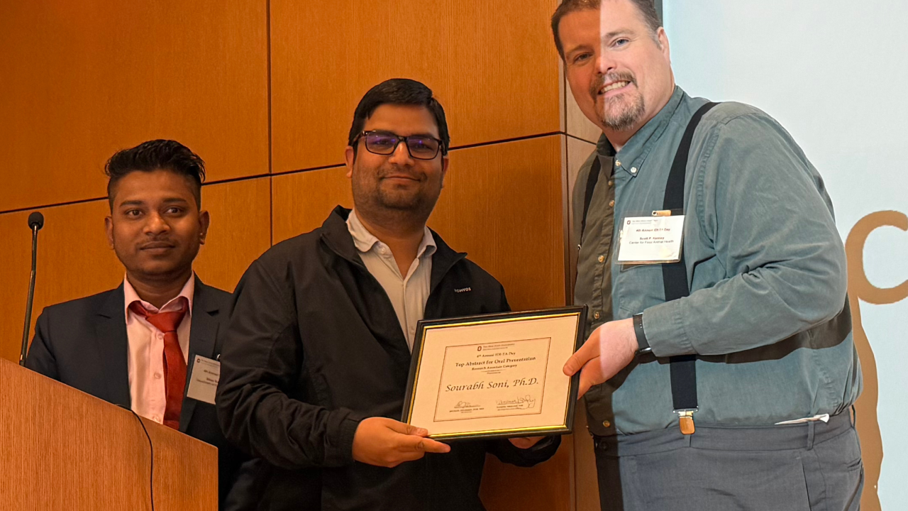 Three individuals stand in front of a wooden wall during an award presentation. One person hands a framed certificate to the recipient in the center, which reads “Sourabh Soni.”