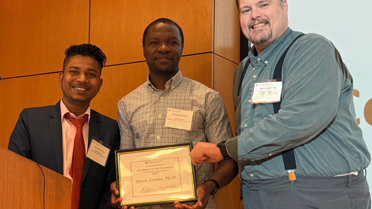 Three individuals stand together during an award presentation. The person in the center holds a framed certificate that reads “Steve Leumi.” The person on the left wears a suit and tie, the center person wears a checkered shirt, and the person on the right wears a blue shirt with suspenders.